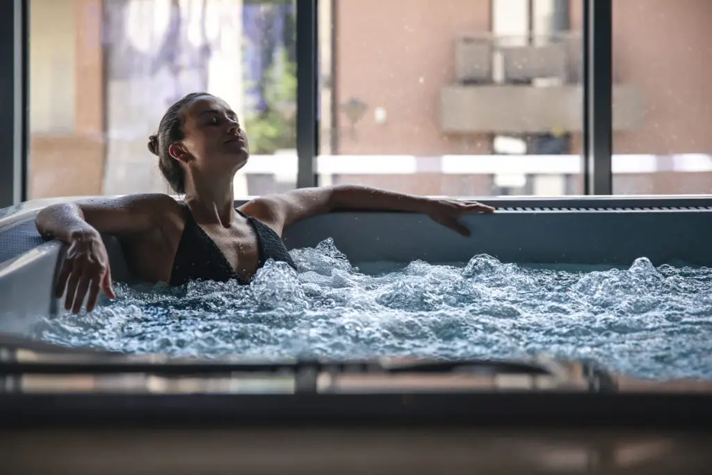 Woman relaxing in a bubbling indoor jacuzzi spa with eyes closed and arms outstretched